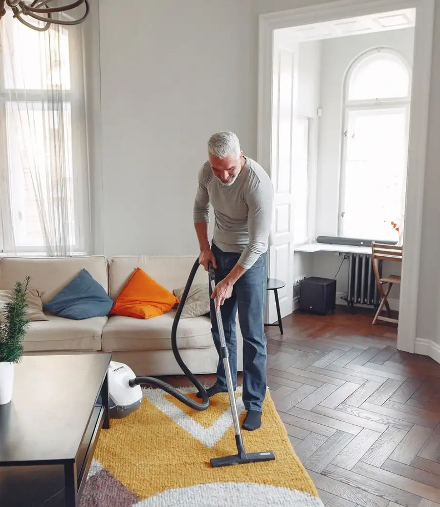 A man vacuuming a patterned rug in a bright living room